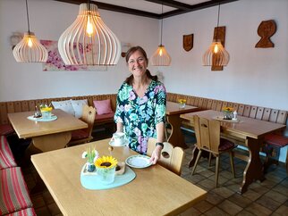 Frühstücksraum im Gästehaus an der Weinstraßenmitte Nicole Ehmer steht lächelnd im hellen Frühstücksraum mit Holztischen, Eckbänken und Holz-Pendelleuchten
