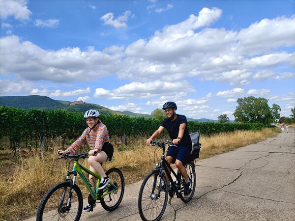 Radtour durch die Weinberge mit Blick auf das Hambacher Schloss Theresa und Daniel Hoefer fahren mit Fahrradhelm auf einem Wirtschaftsweg durch die Weinberge, im Hintergrund das Hambacher Schloss