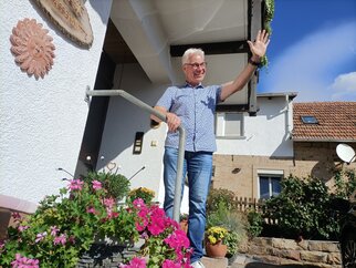 Herzlich willkommen – Gastgeber Matthias Ehmer begrüßt Gäste Matthias Ehmer winkt lachend von der Eingangstreppe des Gästehauses, davor blühende Geranien